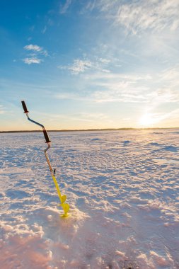 Parlak sarı bıçaklı bir el kumandası, donmuş bir gölde taze bir delikte duruyor. Öğleden sonra ışığı, kar ve buz üzerinde uzun gölgeler bırakıyor..