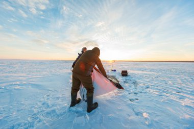İki balıkçı donmuş bir gölün üzerine taşınabilir bir sığınak kurmuş. Uç bir bayrak, kızak, vites kutusu ve ayak izleri hazır takım çalışması gibi güneşin altında duruyor..
