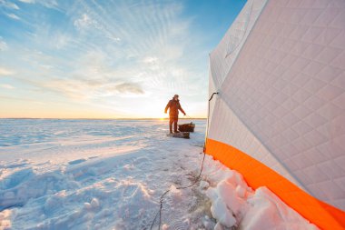 Güneşin doğuşunda donmuş bir gölde duran balık, örtülü turuncu ve beyaz bir sığınak tarafından dişli bir kızak çeker, rüzgarda delikler açarak kış buzunu temizler..