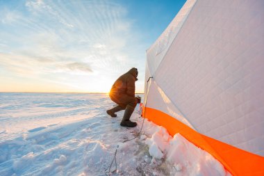 Balıkçı bir bohça deliğin yanında diz çöker, örülmüş beyaz ve turuncu bir çizginin yanında uzun kış gölgeleri olan donmuş bir gölde sığınak oluşturur..
