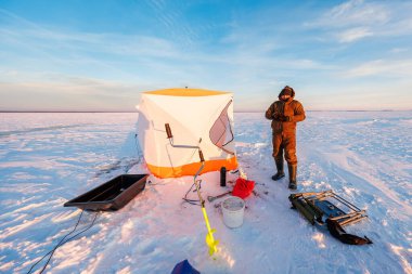 Bir bohça balıkçı donmuş bir gölün üzerinde turuncu ve beyaz bir sığınağın yanında durur. Çapa, kızak, kova, ısıtıcı ve sıcak güneş altında bir sandalye ve uzun gölgeler..