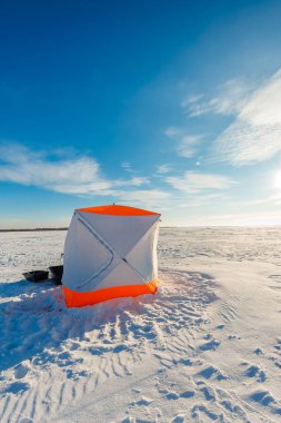 Donmuş bir gölün üzerinde parlak turuncu ve beyaz bir buz sığınağı duruyor. Donmuş bir kızak, delinmiş delikler, uzun gölgeler, rüzgar desenli kar ve görünen erkek çizgileri..