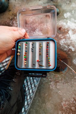 Kışlık botlu bir balıkçı, sert suda delinmiş bir deliğin yanında mikro jigololardan ve tungsten yemlerinden oluşan kompakt bir alet kutusu açar. Kısa bir buz çubuğu çamur ve metal zeminde durur..