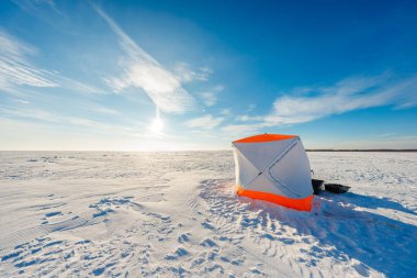 Parlak turuncu ve beyaz renkli bir gecekondu, donmuş bir gölün üzerinde oturur, bir kızak ve teçhizatla birlikte alçak kış güneşi altında, uzun gölgeler altında, ve rüzgar ufka kar oyar..