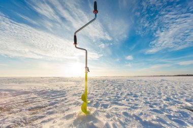 Çift siyah saplı sarı bir el çapası ve turuncu sütyen donmuş bir göl üzerinde durur, kar kabuklu buzun içinde sarmal bir parça, alçak kış güneşi günbatımına yakın uzun gölgeler bırakır.