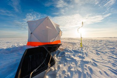 Altıgen bir sığınak, turuncu süslemeli, donmuş bir gölün üzerinde, siyah bir kızak ve sarı bir el açacağının yanında duruyor. Sıcak arka plan ışıkları dalgalı karlar boyunca uzun gölgeler bırakır..