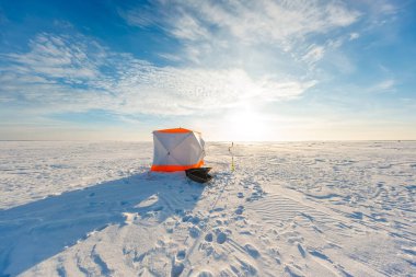 Turuncu ve beyaz bir sığınak donmuş bir göl üzerinde duruyor, bir çapa, siyah bir kızak, ve yumuşak kış güneşi altında bot izleri, geniş ufuk ve nazik bulutlar.