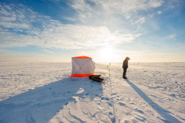 Yalnız bir balıkçı, turuncu budanmış gecekondu ve donmuş bir gölün üzerinde kızak hattını kontrol eder. Bir el çapası taze bir deliğin yanında durur, gün ışığı uzun gölgeler bırakır..