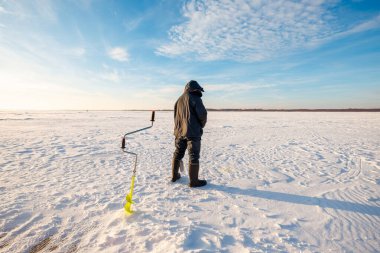 Sarılı bir balıkçı donmuş bir gölün üzerinde durur, sarı bir el çapası, dalgalı kar üzerinde uzun bot gölgeleri, ufukta alçak ağaç çizgisi, geniş açı manzarası, kış öğleden sonra.