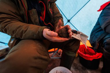 Paketlenmiş bir balıkçı, buzdaki kovanın üzerinde kahverengi bir yem topu şekillendirir. Mavi örtülü bir çadır, kırmızı çizmeli yem kovası, lastik botlar ve gazete şeritleri sıcak ışıkta görülebilir..