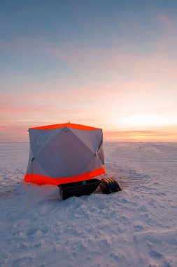 Turuncu buza sahip kompakt altıgen bir buz sığınağı şafak vakti donmuş bir gölün üzerinde, siyah bir kızak ve sarmalanmış teçhizatın yanında, pembe ve altın renkli uzun gölgeler altında sürüklenir..
