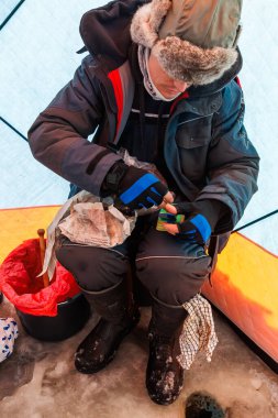 Paketlenmiş bir balıkçı örtülü bir sığınakta oturur, matkapla delinmiş bir buz deliğinin yanında yem ve teçhizat hazırlar ve ıslak buzun üzerinde kırmızı bir torba ile bir kova, yakın görüş odak noktaları