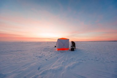 Tek bir balıkçı, donmuş bir ovada turuncu ve beyaz bir gecekondunun yanında bir kızak ve kova hazırlar. Yumuşak pembe ışık buzu kaplarken ayak izleri karları aşıyor..