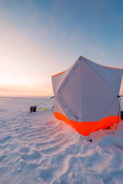 Güneş doğarken rüzgârlı, donmuş bir gölün üzerinde duran turuncu buza budanmış bir gecekondu, yanında bir çubuk, kova ve çapa ile. Uzun gölgeler ve pastel ışık sahneyi tanımlar.