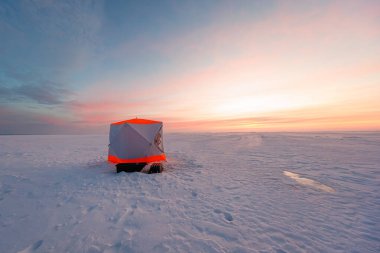 Parlak turuncu buzullu bir buz sığınağı güneşin doğuşunda donmuş bir gölün üzerinde duruyor. Zayıf bot izleri ve yarım gömülü bir kızak yakınlarda duruyor. Yumuşak düşük ışık uzun gölgeler bırakır.
