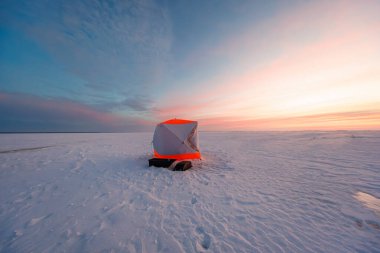 İki siyah kızak ve soluk bot izleriyle donmuş bir gölün üzerinde turuncu ve gri bir barınak beliriyor. Yumuşak pastel ışık geniş bulut bantları altında geç bir gün olduğunu gösteriyor..