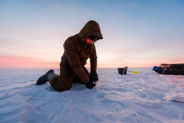 Paketlenmiş bir balıkçı, şafak vakti bir buz deliğinin önünde diz çöker. Siyah bir kova, sarı kepçe ve kızak alacakaranlık altında dalgalı karların üzerinde oturuyor..