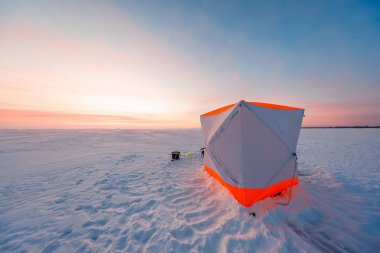 Alacakaranlıkta donmuş bir gölün üzerinde turuncu budanmış bir sığınak duruyor. Rüzgârlı kar ve uzun gölgelerin içinde dinlenen bir çubuk, kova ve kızak..