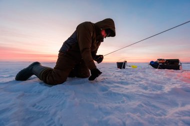 Yalnız bir balıkçı şafak vakti donmuş bir gölde, delinmiş bir delikte diz çöker, elinde kısa bir çubuk, yanında kızak ve kova, soluk görünür, dalgalı kar ve botların üzerinde pastel bir ışık
