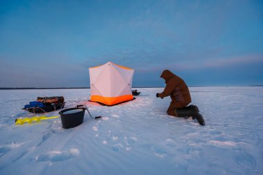 Solo bir balıkçı beyaz ve turuncu bir barınağın yanında diz çöker, buza doğru bir çizgi çeker, sarı bir çengelli, siyah bir kova, ve alacakaranlıkta rüzgarda karla işaretlenmiş teçhizat kızağı.