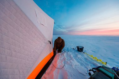 Paketlenmiş bir balıkçı, yalıtımlı bir sığınakta gün batımında adam kuyruğu oluşturur. Sarı bir el çapası, bir kızak ve bir kova kış alacakaranlıklarında donmuş bir alanda dinleniyor..
