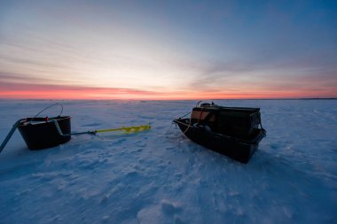 Teçhizatla dolu iki siyah kızak kış günbatımında kar işaretli rüzgarın üzerinde oturuyorlar. Parlak sarı bir el çapası onları birbirine bağlıyor. Geniş alçak görüş, büyük donmuş bir gölü gösteriyor..