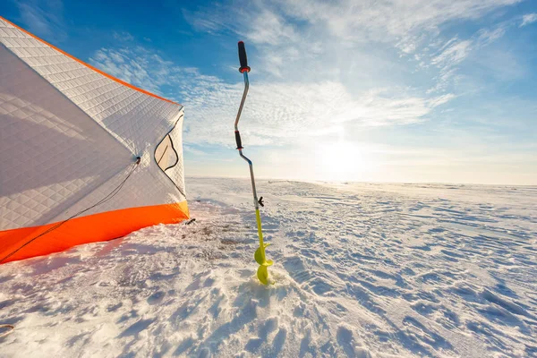 Turuncu ve beyaz yorgan kaplı bir sığınak donmuş bir gölün üzerinde yeşil bir çengelin yanında duruyor. Uzun gölgeler kışın düşük bir güneşe kadar uzanıyor..