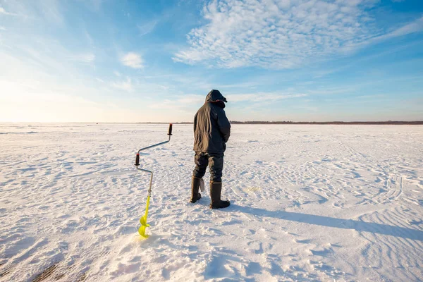 Sarılı bir balıkçı donmuş bir gölün üzerinde durur, sarı bir el çapası, dalgalı kar üzerinde uzun bot gölgeleri, ufukta alçak ağaç çizgisi, geniş açı manzarası, kış öğleden sonra.