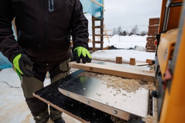 Neon işlemeli eldivenler bir tahtaya taşınabilir kalınlık planöründen uçarken rehberlik eder. Kar, kereste yığını ve geçici çerçeve modern bir evde soğuk sezon çalışmalarını gösteriyor..