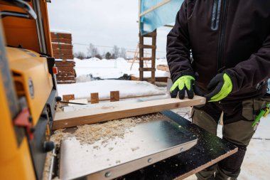 Neon eldivenli bir marangoz portatif kalınlıkta bir tahtayı besler. Talaşlar, muşambalarla ve iskelelerle kaplı karlı bir iş yerindeki metal bir taşıma masasına saçılır..