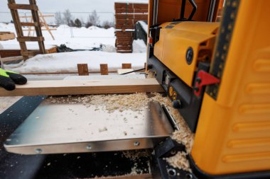 Parlak sarı bir planör, eldivenli bir işçinin rehberlik ettiği ahşap bir tahtayı buduyor. Taze talaşlar masaları kaplar. Kar, kereste yığını ve çit, kış çerçevesini işaret ediyor..