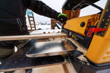 Kışlık giysili bir marangoz, açık hava ahşap çerçeve saçılmış yeni talaşlar, iskeleler ve istiflenmiş keresteler aktif bir işi işaret ediyor..