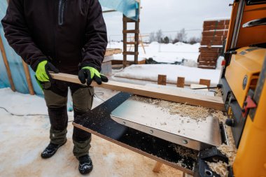 Yalıtımlı ekipman işçisi portatif kalınlıkta uzun bir tahtayı besliyor, talaşlar ayak altında, yalıtım torbaları ve yakındaki kerestelerle birlikte, dış beslenme masasına saçılırken..