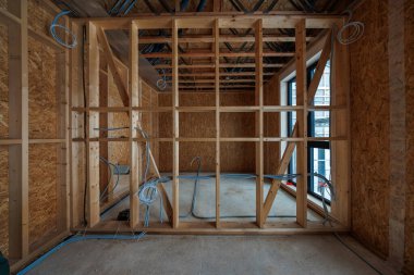 İç ahşap çerçeveli oda açıkta duran çiviler, OSB kılıfları, elektrik kabloları, çapraz destekleme, açık kirişlerdeki kanallar ve yumuşak doğal ışıkta uzun bir çift pencere gösteriyor..