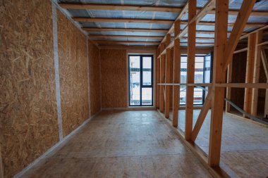 İç görünüm çerçeve, OSB, buhar kontrol zarları ve uzun pencereleri gösteriyor. İşçiler çerçeve ve hava mühürü, servis boşlukları kablolama ve ahşap fiber gevşek dolgu yalıtımı bekliyor.