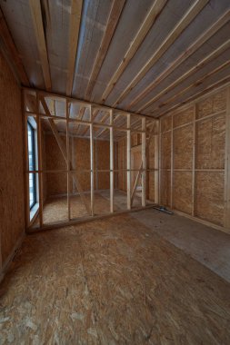 İnşaat altındaki modern ahşap çerçeve, çivili duvarlar, OSB kılıfları, açıkta kalan kirişler ve geçirgen buhar zarını gösteriyor. Dar bir pencere ve süpürge kapalı alanda görülebilir..