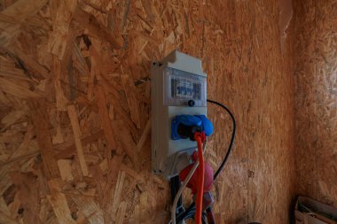 Şeffaf kapaklı kompakt sigorta kutusu ve mavi soketler OSB duvarında turuncu ve siyah kablolar taşır, sıcak yön ışığı ve erken faz kurulumunu göster.