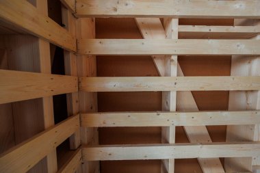 İç tavan çerçevesi, soluk ladin kirişleri, çapraz teller ve OSB panellerini gösteriyor. Ilık yayılmış ışık testere izleri, düğümler, pilot delikleri ve metal bağlayıcıları ortaya çıkarır..