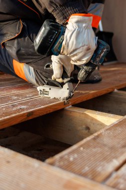 Yakından bakınca matkap ve yapboz kullanan eldiven işçisi görülüyor. Tahta talaşı mevcut. Ilık ışık tahılı ve açık altyapıyı ortaya çıkarır, teknik ve güvenliği vurgular..