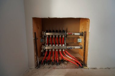 Kırmızı PEX döngüleri ve metal başlıklarla donatılmış hidronik manifoldlar, etiketli kapaklar, kapatma vanaları ve gündüz işleri sırasında çift katlı katmanlı kapaklı kapaklı kapakçıklarla kabaca duvar açıklığında otururlar.