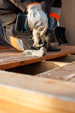 Tradesperson kereste kirişleri üzerine diz çöker, kablosuz matkap ve klemple ahşap bir tahtayı hizalayıp tutturur. Buz, kalasların tozunu alır. Soğuk mevsim günışığını işaret eder..