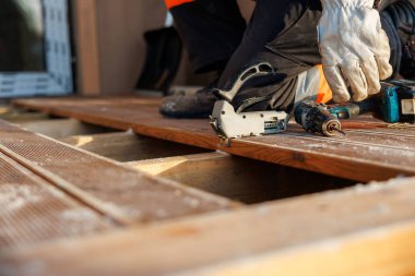 Eldivenli bir işçi ahşap kirişlerin üzerine diz çöker, matkap sürücüsü ve bağlanma aleti olarak ahşap bileklikleri hizalayıp yakınlarda oturur. Geç saatlere kadar gün ışığı talaş ve tahılın altını çizer.