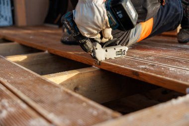 Eldiven işçisi oyuk ahşap tahtaları kablosuz matkapla ve gündüz şarjörleriyle kirişlere sabitliyor. Dar açı yakın çekim, talaş, vida kafalar ve hizalı altyapı gösteriyor.