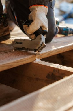 Beyaz eldivenli bir zanaatkar, güverte tahtasına metal çivi tabancası takarak gün ışığında açık bir alanda kirişlere bağlanır. Dikiş yeri boyunca talaş ve sığ bir alan derinliği vardır..