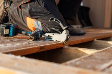 Güvenlik botu giymiş bir işçi bitmemiş ahşap bir güvertede diz çöker ve aletler yakınlarda dinlenirken kirişlere tahtaları sabitler. Sıcak gün ışığı ve sığ alan derinliği zanaatı vurguluyor.