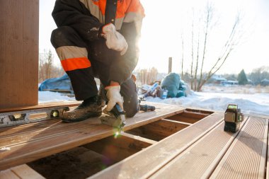 Turuncu ve siyah ceketli bir işçi ahşap bir güvertede diz çöker, çivi, ruh seviyesi, kablosuz matkap kullanır ve güneş ışığı çıplak ağaçların ve karın içinde parlarken yeşil lazer seviyesi.