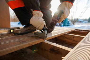 Eldivenli bir işçi, döşemeyi matkap ve zımba tabancasıyla kirişe bağlıyor. Yerde kar var ve alçak öğleden sonra ışığı bağlayıcılarda buza sıcaklık katar..