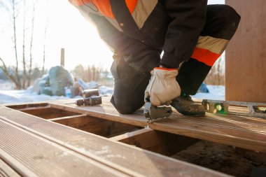 Yalıtımlı teçhizatlı bir işçi, kışın ışık altında ruhsal düzeyi ve kablosuz matkabı olan bir çivi kullanarak kereste iskeletine birleştirme tahtalarını bağlar..