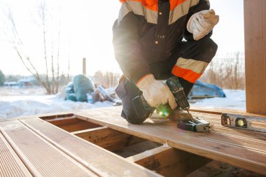 Dışarıdaki bir traDesperson sökülmüş ahşap bir güvertede diz çöker. Kablosuz matkabı ruh seviyesi ve mezura olarak kullanır. Kış gündoğumunda kar ve çıplak ağaçlarla birlikte yakınlarda dinlenir..