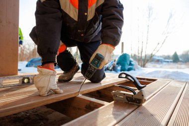 Güverte yükleyicisi kirişlerin üzerine diz çöker, kablosuz matkapla kompozit tahtalara vida sokar. Arkasında kar var, aletler yakınlarda. Sıcak öğleden sonra ışık işaretleri soğuk sezon çalışmaları.